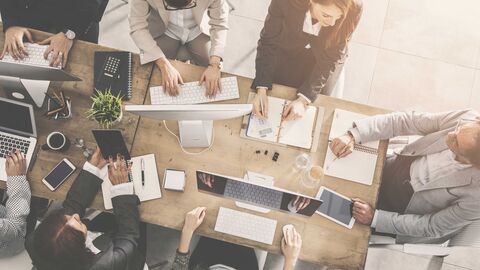 Zusammenarbeit Mehrere Personen sitzen an einem Tisch und haben verschiedene Unterlagen und Laptops vor sich stehen. Sie unterhalten sich und arbeiten gemeinsam an einem Projekt